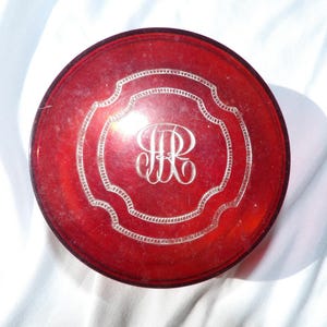 May include: A circular, deep red glass powder jar featuring a translucent finish. The lid is decorated with an etched, scalloped border and a central, ornate monogram consisting of the intertwined letters HR in a white, engraved script style.