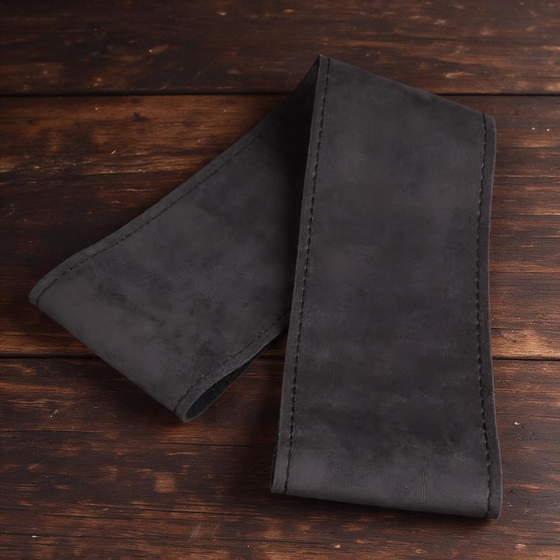 Pu&ograve; includere: Una lunga striscia rettangolare di pelle grigio antracite scuro con una texture opaca simile al camoscio. I bordi della striscia di pelle sono rifiniti con una cucitura scura e coerente che corre lungo l'intero perimetro del pezzo.
