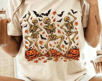 Può includere: Una t-shirt color crema con una stampa grafica di tre scheletri gialli che danzano tra zucche arancioni, foglie autunnali sparse e pipistrelli neri. Gli scheletri sono posizionati in varie pose con zucche ai loro piedi.