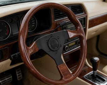 Può includere: Un volante per auto con corona in legno lucido e mozzo centrale nero opaco a tre razze. L'interno presenta un cruscotto beige, finiture in legno, una leva del cambio nera e un quadro strumenti con indicatori analogici.
