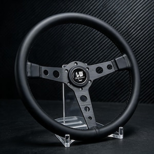 May include: A black steering wheel featuring a circular rim and three brushed metal spokes with circular cutouts. The centre horn button displays the text LW Performance in a white script font. The design includes a black finish throughout.