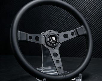 Può includere: Un volante nero caratterizzato da un cerchio circolare e tre razze in metallo spazzolato con fori circolari. Il pulsante centrale del clacson riporta la scritta LW Performance in un carattere corsivo bianco. Il design presenta una finitura nera.