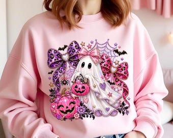 Può includere: Una felpa girocollo rosa chiaro con una grafica centrale raffigurante un fantasma bianco con un fiocco rosa, che tiene in mano un secchiello a forma di zucca rosa. Il design include fiocchi leopardati viola e rosa, ragnatele, pipistrelli e stelle.