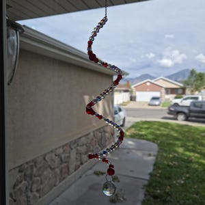 May include: A spiral-shaped suncatcher hanging from a thin metal chain, constructed with alternating clear iridescent beads and solid red beads, terminating in a single faceted clear crystal drop at the bottom of the twisted wire structure.