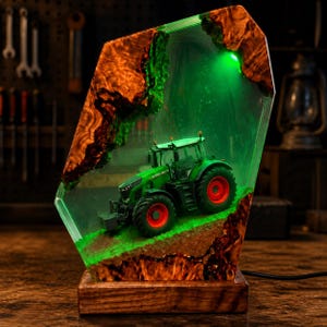 May include: A decorative lamp featuring a miniature green tractor model encased in clear resin with natural wood edges. The piece emits a bright green light from within, illuminating the tractor and the textured brown base inside the resin structure.