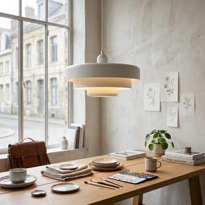May include: A matte white tiered pendant light fixture with three concentric circular shades suspended from a single cord, featuring a clean, layered design that directs light downward, creating a soft illumination in a room.