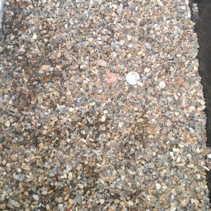 Puede incluir: Una colección de pequeñas piedras de grava multicolor en tonos gris, bronceado y marrón. Una moneda de color cobre y una moneda de color plata están colocadas en el centro de las piedras para proporcionar una escala visual del tamaño de la grava.