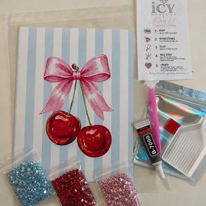 May include: A DIY rhinestone art kit featuring a print of two red cherries with a pink bow on a light blue striped background. Includes three bags of rhinestones in light blue, red, and pink, a tube of B-7000 glue, a pink applicator pen, a white tray, and instructions.