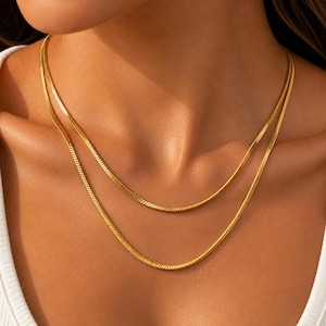 May include: A layered gold-tone snake chain necklace featuring two strands of varying lengths. The chains have a smooth, flat, and flexible design with a polished metallic finish that reflects light, resting against the collarbone area.