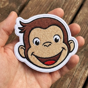 May include: An embroidered patch featuring the face of a cartoon monkey with brown fur, a tan face, and a wide smile. The character has large black eyes, a small nose, and a red tongue visible inside the mouth. The patch has a thick white border.