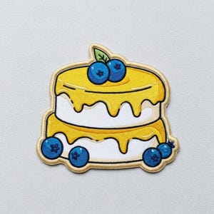 May include: An embroidered patch shaped like a two-tiered cake with white frosting, yellow icing drips, and blue blueberries. The design features a green leaf on top and a light tan border, creating a detailed, colorful, and textured fabric accessory.