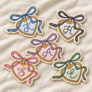 May include: Five heart-shaped embroidered patches with gold borders, each featuring a different pastel center color, a tied ribbon at the top, and a single cursive letter: K in blue, H in grey, A in purple, S in pink, and E in green.