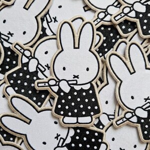 May include: A collection of embroidered patches featuring a white rabbit character with long ears, black eyes, and a cross-shaped mouth. The rabbit wears a black dress with white polka dots and is shown playing a small flute instrument.