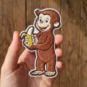 May include: An embroidered patch featuring a brown monkey with a light tan face, hands, and feet, holding a yellow banana. The design is outlined in white thread and depicts the character standing upright with a smiling expression and wide eyes.