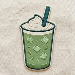May include: An embroidered patch shaped like a cup of iced matcha latte. The design features a light green liquid with several square ice cubes, topped with a swirl of cream and a dark green straw. The entire patch has a dark green border.