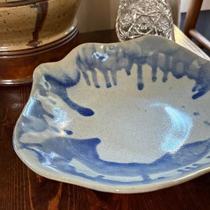 May include: A shallow ceramic bowl with an irregular, wavy rim. The interior surface features a light, speckled off-white base glaze with vibrant blue glaze drips running from the edges toward the center, creating a distinct contrast in color.