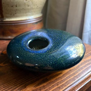 May include: A low, disc-shaped ceramic flower frog vase with a glossy, speckled glaze in deep shades of blue and green. The circular vessel features a central opening that reveals a metal pin holder inside for arranging stems securely.