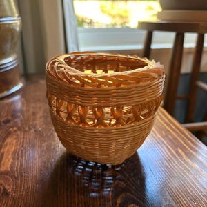May include: A small, round, light brown woven basket with a smooth, natural finish. The basket features a tight, solid weave at the base that transitions into an open, decorative lattice pattern near the top rim, which is finished with a braided edge.
