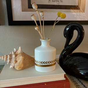May include: A white ceramic bud vase with a gold laurel leaf band, holding dried poppy pods and yellow billy balls. The vase sits on a stack of books next to a large tan seashell and a black ceramic swan figurine on a flat surface.