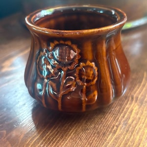 May include: A small, round, brown glazed ceramic pot featuring a relief design of three sunflowers on the exterior. The vessel has a wide, open mouth and a slightly flared rim, with vertical ribbed detailing around the body of the container.