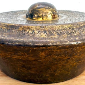 May include: A round, dark bronze-coloured metal gong with a central raised boss. The surface features a weathered, textured patina with gold-toned highlights and intricate, etched geometric patterns encircling the top edge and the central dome.