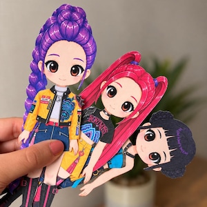 Op de afbeelding: Drie uitgestanste papieren poppen met anime-stijl personages met grote ogen. Eén heeft paars gevlochten haar, een geel jasje en een blauwe rok. Een andere heeft roze staartjes en een zwart shirt met de tekst WONT MISS. De derde heeft zwart haar.