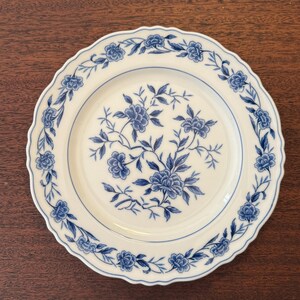 May include: A round white ceramic plate with a scalloped edge, featuring a blue floral pattern. The center of the plate displays a detailed blue botanical illustration of flowers and leaves, while the rim is decorated with a repeating floral border.