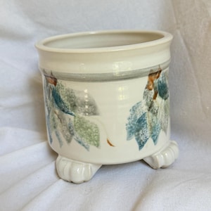 May include: A cream-colored ceramic planter with a cylindrical body and three small, rounded feet. The exterior features a hand-painted design of blue and green leaves with brown accents, and a horizontal gray band encircling the upper rim.