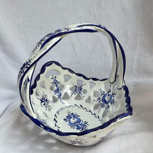 May include: A white ceramic basket with a blue floral pattern and a lattice-style openwork design. The basket features a curved handle, a scalloped rim, and hand-painted blue flower motifs on the interior base and along the exterior handle.