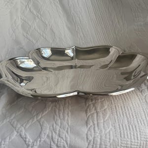 May include: A polished silver-tone metal serving dish with a distinct four-lobed, clover-like shape. The bowl features smooth, reflective surfaces and curved, scalloped edges that create a shallow, wide basin for holding various small items.