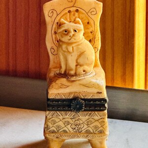 May include: A cream-colored carved trinket box shaped like a chair with a cat figurine seated on the seat. The base features etched geometric patterns and a central flower design, while a dark metal hinge with a floral clasp secures the lid.