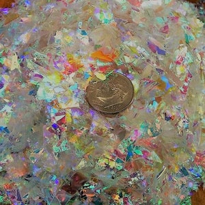 May include: A mound of iridescent, translucent, and colourful glitter flakes. The flakes are scattered on a wooden surface, with a US dime coin for size reference. The glitter reflects light, creating a rainbow effect.