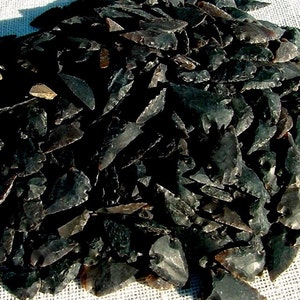 May include: A pile of dark obsidian arrowheads, each with a sharp point and a notched base. The arrowheads vary in size and shape, with a mix of smooth and textured surfaces. The color ranges from black to dark gray, with some showing hints of brown.