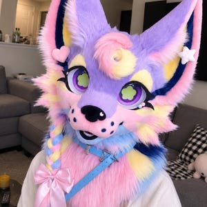May include: A plush animal head mask featuring pastel purple, pink, yellow, and blue faux fur. The mask has large purple eyes with flower-shaped pupils, a black nose, a braided section with a pink satin bow, and a light blue collar with a star charm.