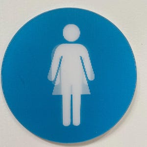 May include: A round, light blue sign with a white pictogram of a person. The person is depicted with a head, shoulders, arms, and a skirt-like shape, indicating a female restroom. The sign is mounted on a white wall.