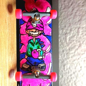 Pode incluir: Um fingerboard com uma ilustração de personagem de desenho animado com moletom verde, calça azul e gorro roxo. O deck tem um design de fundo rosa e azul vibrante, equipado com trucks de metal prateado e rodas de plástico rosa.
