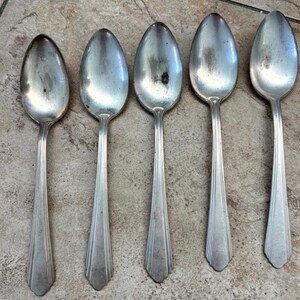 Puede incluir: Cinco cucharas de metal plateado dispuestas en fila. Cada cuchara presenta un cuenco ovalado y un mango con tres estrías verticales que se estrechan hacia un extremo puntiagudo. Las superficies metálicas muestran desgaste y manchas oscuras.