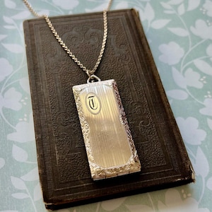 May include: A silver rectangular pendant necklace with a chain, resting on a dark brown book. The pendant has a vertical striped design with an oval containing the letter "T". The pendant is framed with an ornate floral design.