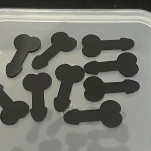 May include: A collection of small, flat, black phallic-shaped cutouts arranged inside a clear, rectangular plastic storage container. The individual pieces are uniform in color and shape, featuring a rounded top section and a tapered base.