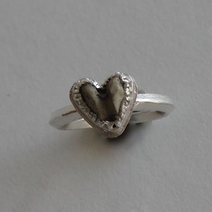 May include: A silver ring with a heart-shaped stone set in the center. The stone is a dark gray color and has a textured surface.