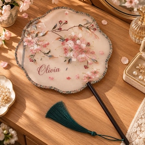 Peut inclure: Un éventail à main translucide aux bords festonnés, orné de broderies de fleurs de cerisier roses et du prénom Olivia inscrit en lettres rouges. L'éventail possède un manche en bois sombre et un long pompon vert foncé attaché à la base.