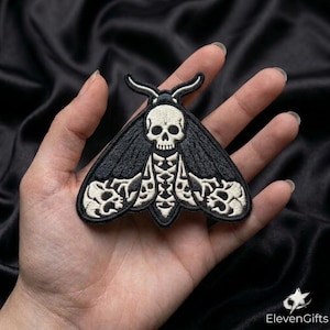 May include: An embroidered patch in the shape of a moth with black wings and a cream-colored skull at the center. The wings feature intricate cream-colored skull patterns along the edges, and the moth has two curved antennae at the top.