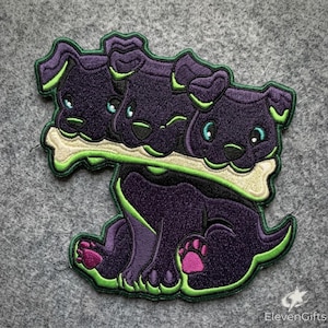 May include: An embroidered patch featuring a three-headed puppy character with dark purple fur, bright green accents, and light blue eyes. The creature holds a large, cream-colored bone in its mouths. The design includes pink paw pads.