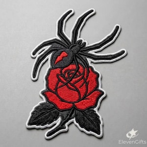 May include: An embroidered patch featuring a black spider with a red hourglass marking on its abdomen, positioned above a vibrant red rose with dark black leaves. The entire design is outlined with a thick white border for clear definition.