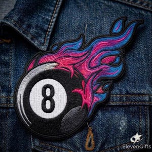 May include: An embroidered patch featuring a black billiard ball with a white circular center containing the number eight, surrounded by stylized flames in shades of vibrant pink, magenta, and blue, with a black border and a needle at the top.