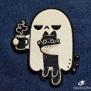 May include: An embroidered patch featuring a cream-colored ghost holding a black mug with rising steam in one hand and hugging a small black cat with white eyes in the other. The design has a thick black border and is set against a blue denim fabric.