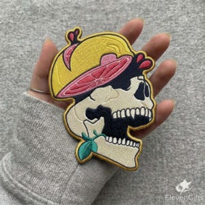 May include: An embroidered patch featuring a cream-colored human skull wearing a yellow citrus slice hat. The skull has dark navy blue shading around the eye socket and jaw, with a small green leaf accent near the chin and pink fruit details.