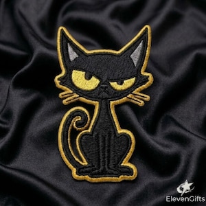 May include: An embroidered patch featuring a black cat with a grumpy expression, large yellow eyes, and pointed ears. The cat is depicted in a sitting position with a curled tail, outlined in a thick gold thread border against a black fabric.