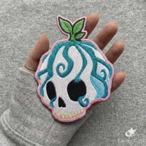 May include: An embroidered patch featuring a white skull with black eye sockets and a nose cavity. The skull is adorned with swirling teal blue vines and topped with two green leaves. The entire design is outlined with a light pink stitched border.