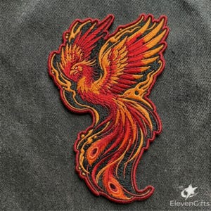 May include: An embroidered patch featuring a phoenix in flight with vibrant red, orange, and yellow thread colors. The mythical bird is depicted with outstretched wings and a long, flowing tail, outlined in black thread against a dark gray fabric.